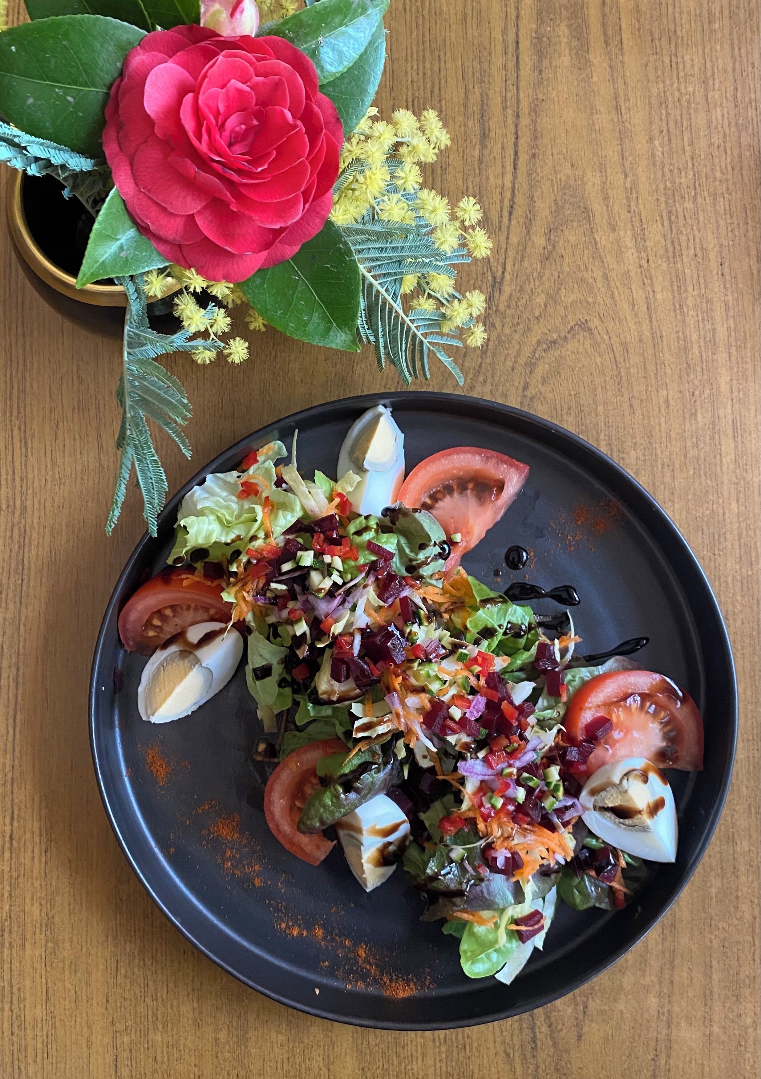 A plate of salad with tomatoes, lettuce, and other vegetables, accompanied by a red rose and yellow flowers.
