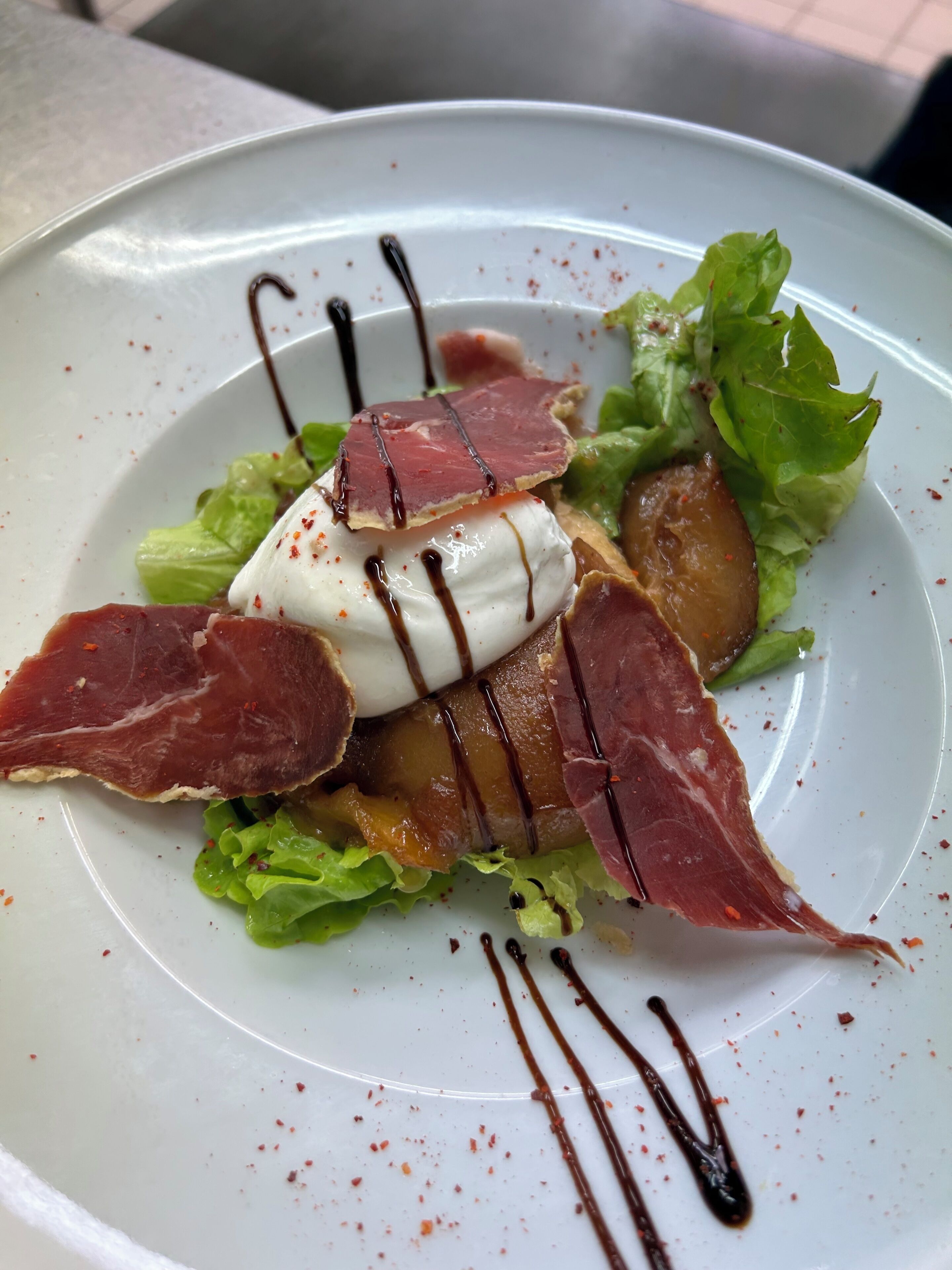 Un plato con ensalada, mozzarella, jamón serrano y un chorrito de salsa.