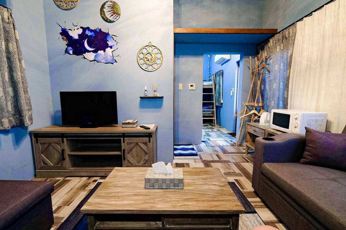 Un salon avec un téléviseur, une table basse en bois et un canapé. Un mur est décoré de différents objets, et le sol est recouvert d’un motif.