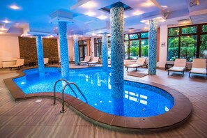 An indoor swimming pool with tiled columns, lounge chairs, and a view of greenery outside. - Orbilux (Bansko)