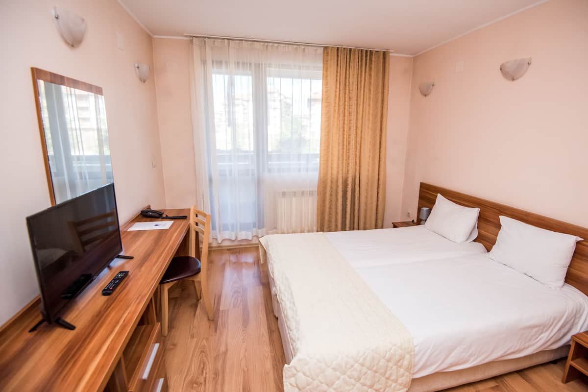 A hotel room with a wooden desk, a TV, a bed with white bedding, a chair, and a window with curtains.