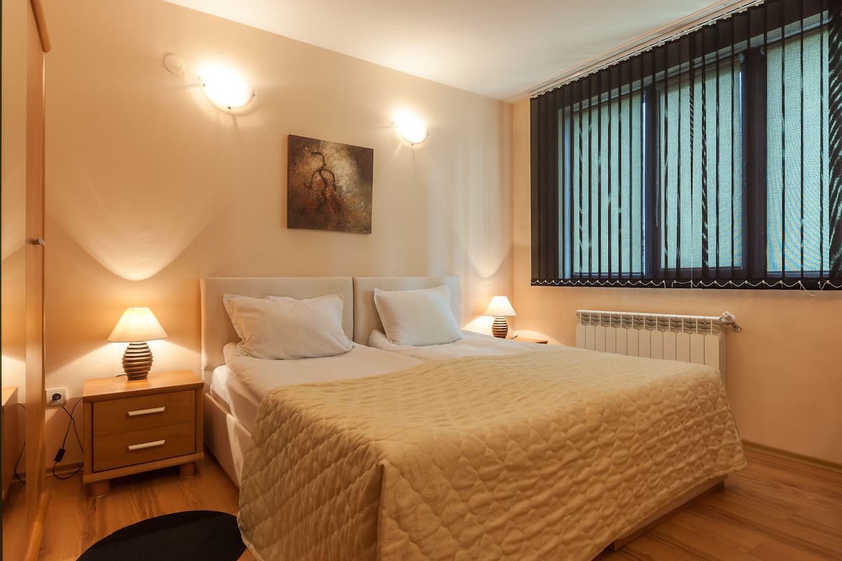 A bedroom with a bed, bedside lamps, a nightstand, a radiator, and a window with blinds.
