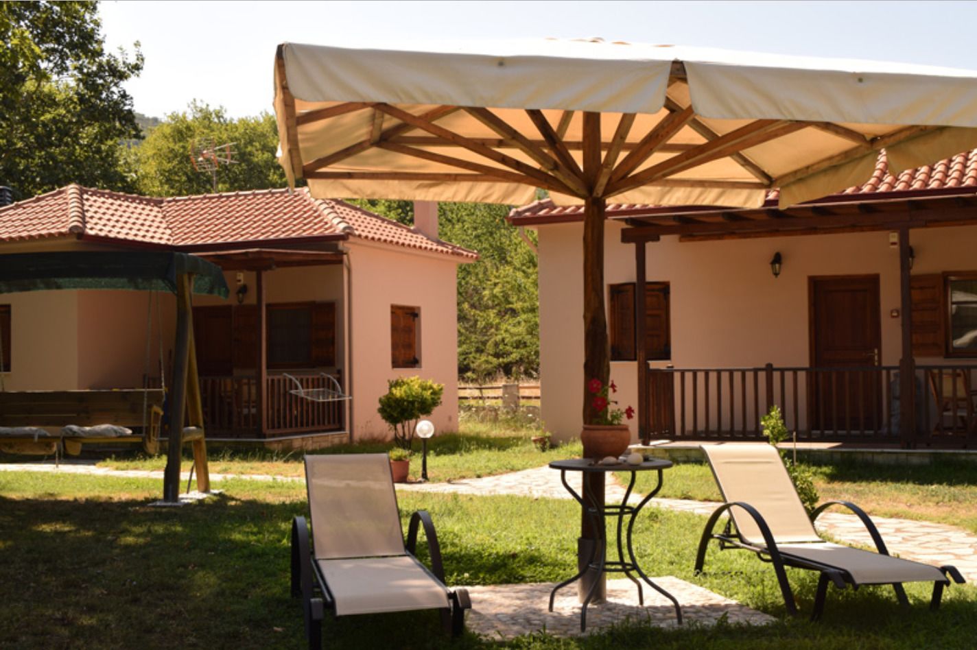 A patio area with two lounge chairs and a table under a large umbrella, surrounded by a small building with a tiled roof and a covered seating area.