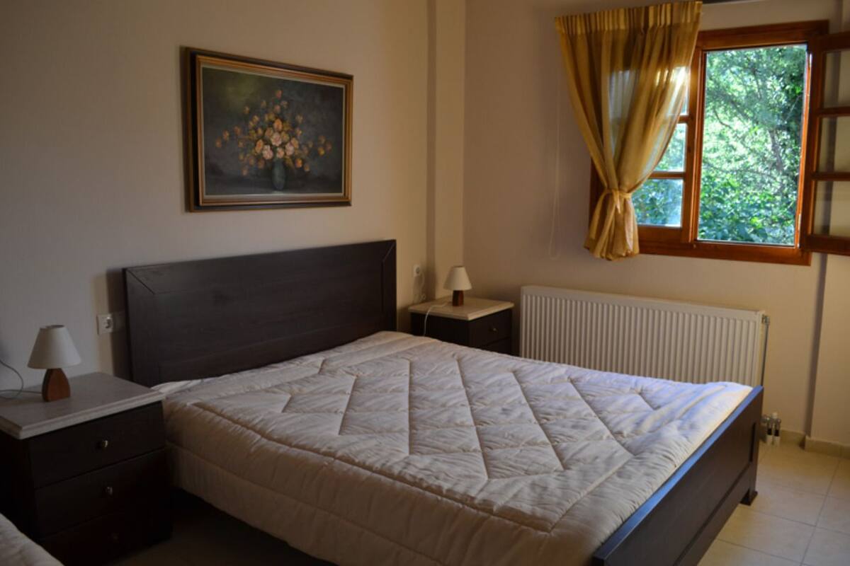 A bedroom with a bed, bedside tables, a nightstand, a painting, a window with curtains, and a radiator.