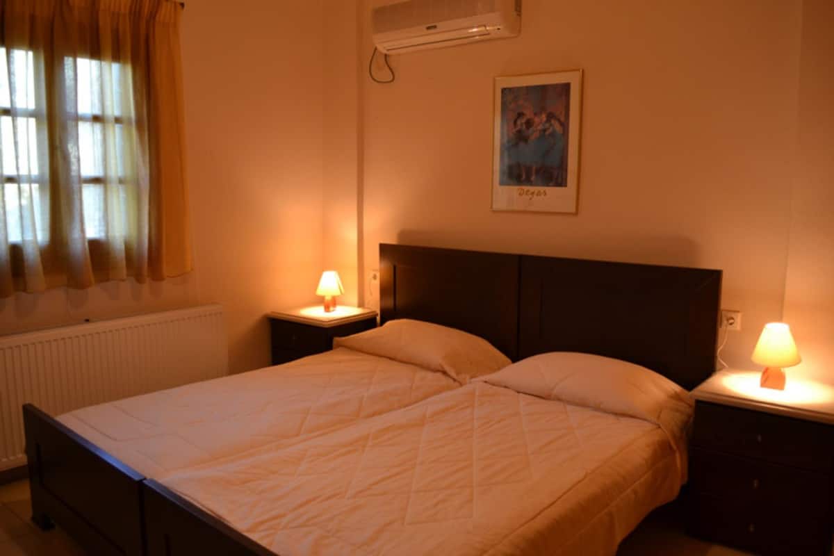 A double bed with two nightstands, each with a lamp, in a room with a radiator, a window with curtains, and a framed picture on the wall.