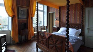 A four-poster bed with a wooden headboard, a television on a small table, and a chair in a room with patterned wallpaper and curtains. - Le Château de la Vierge (Bures-sur-Yvette)