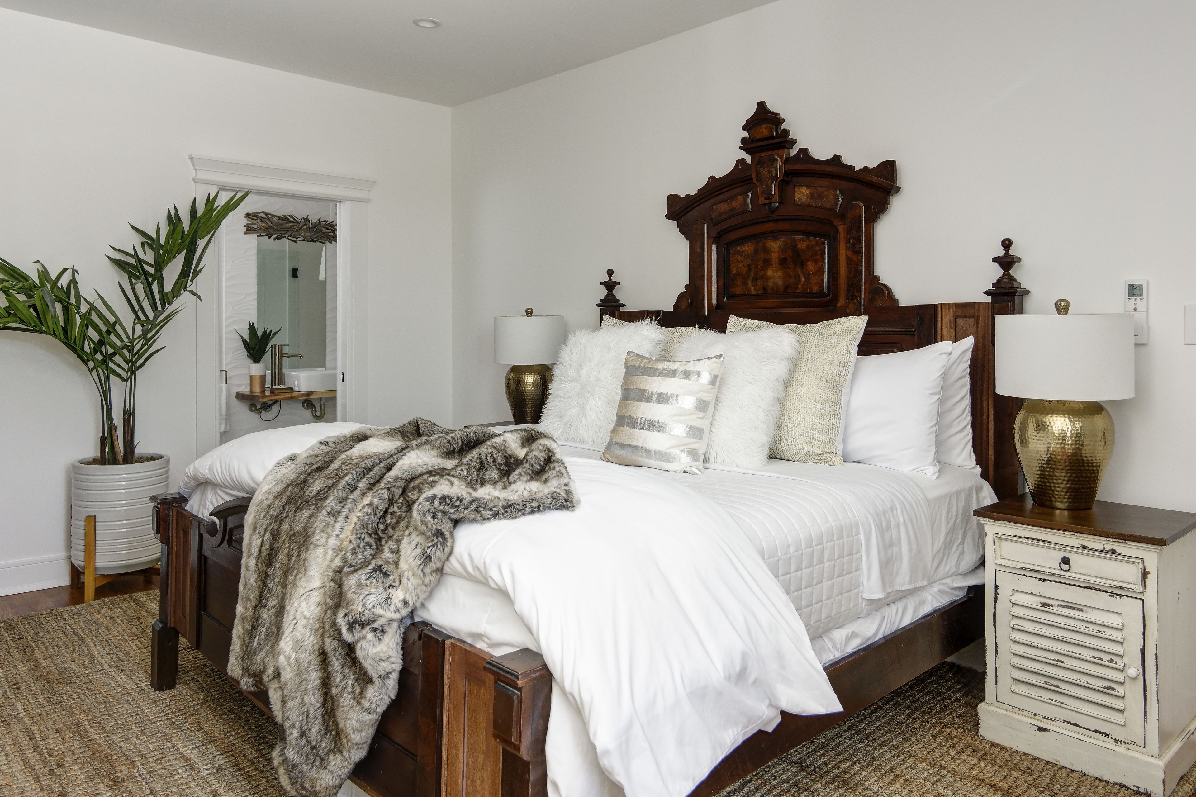 A bedroom with a large bed, two bedside tables, a nightstand, a potted plant, and a mirror.