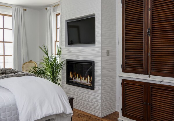 A bedroom with a bed, a fireplace, a television, and wooden shutters.