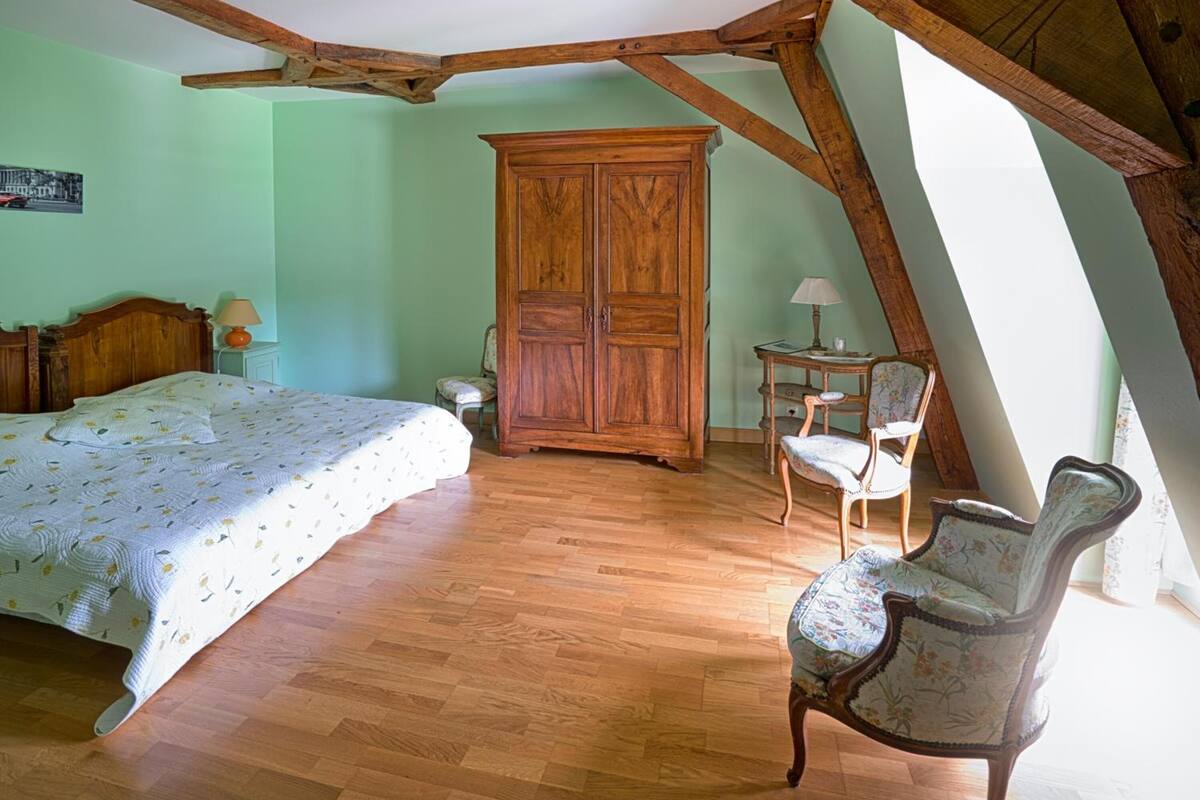 A bedroom with a wooden bed, a wooden wardrobe, a chair, and a small table with a lamp.