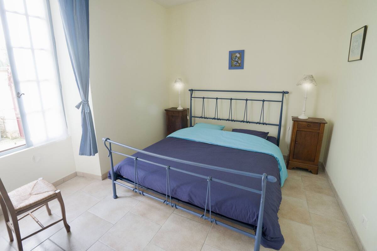 A bedroom with a metal bed frame, a blue and purple bedspread, two bedside lamps, a small cabinet, and a chair.