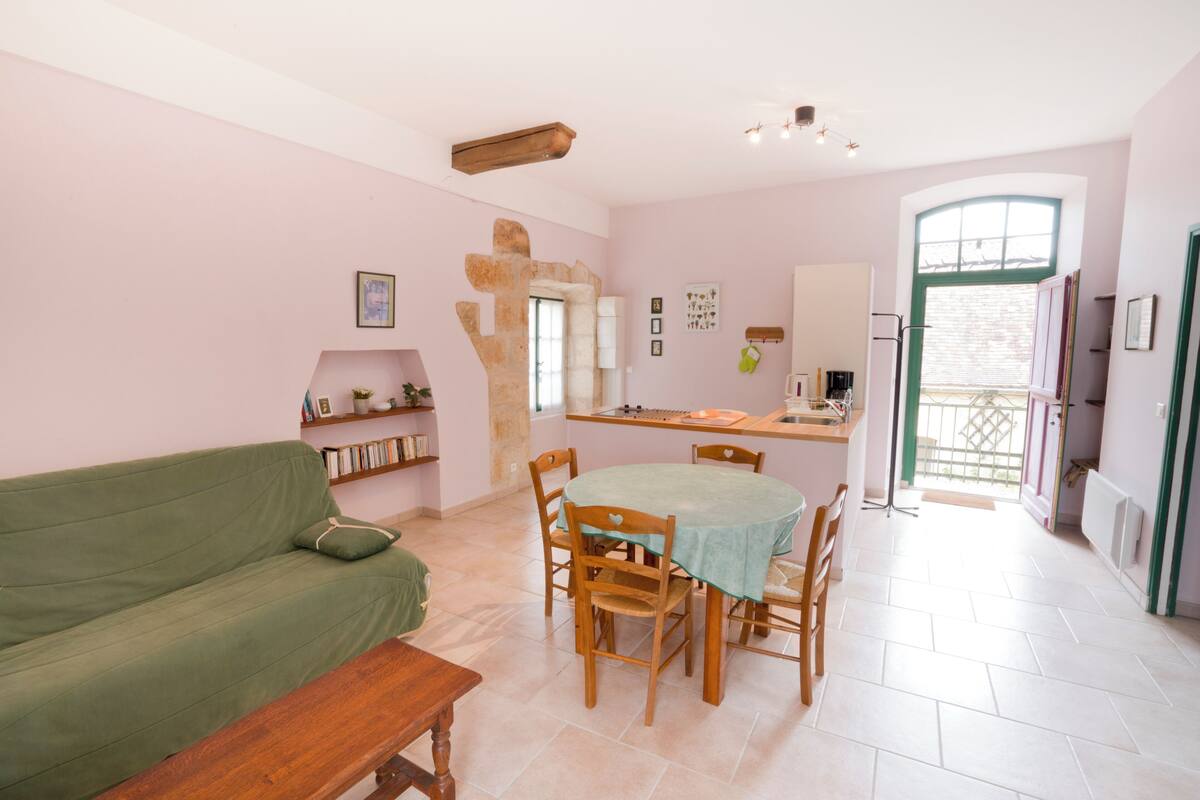 A cozy living room with a green sofa, wooden dining table, and chairs. A kitchen area with a window and a door leading outside is visible.
