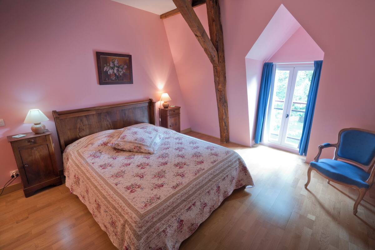A bedroom with a wooden bed, a nightstand with a lamp, a chair, and a window with curtains.