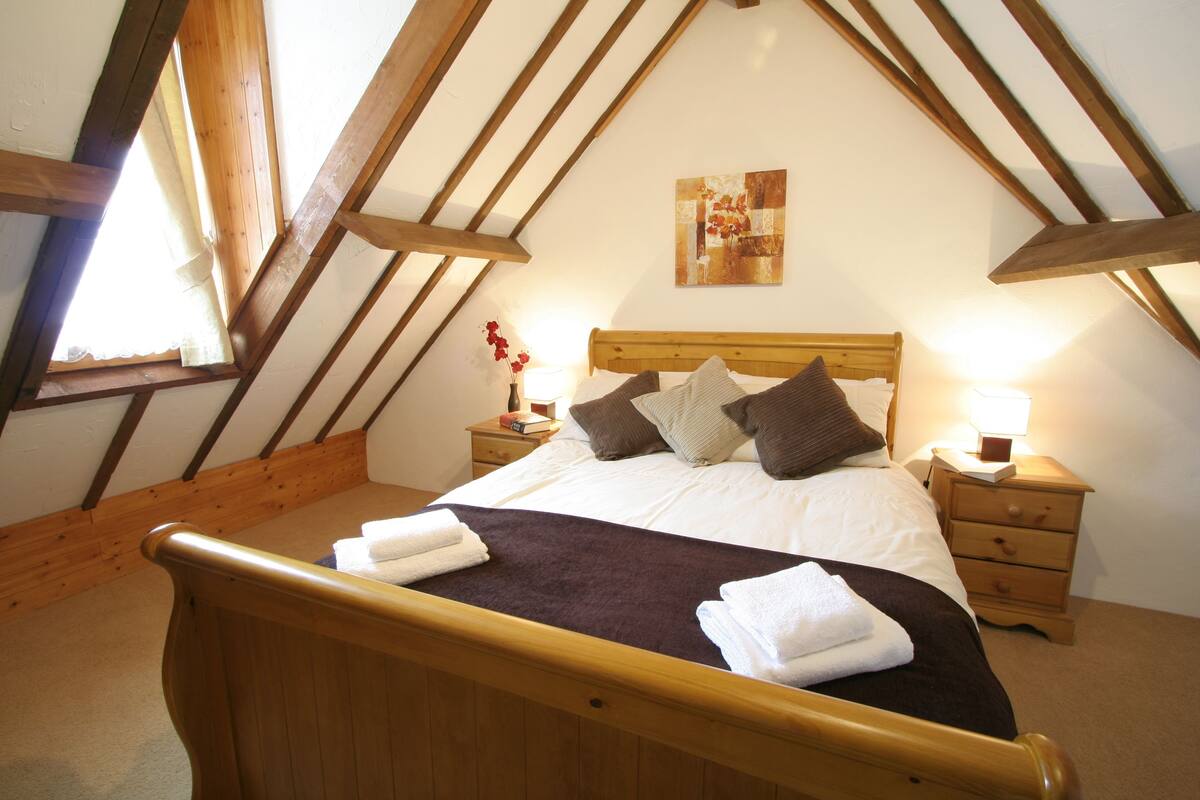 A bedroom with a wooden ceiling, a bed with white linens, a nightstand with a lamp, and a painting on the wall.