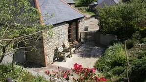A stone house with a tiled roof, a patio with wooden furniture, and a garden with flowering plants.