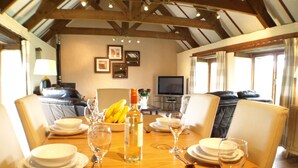 A dining area with a wooden table set for four, surrounded by leather chairs. A television is mounted on the wall, and there is a sofa in the background. - Polean Farm Cottages (Looe)
