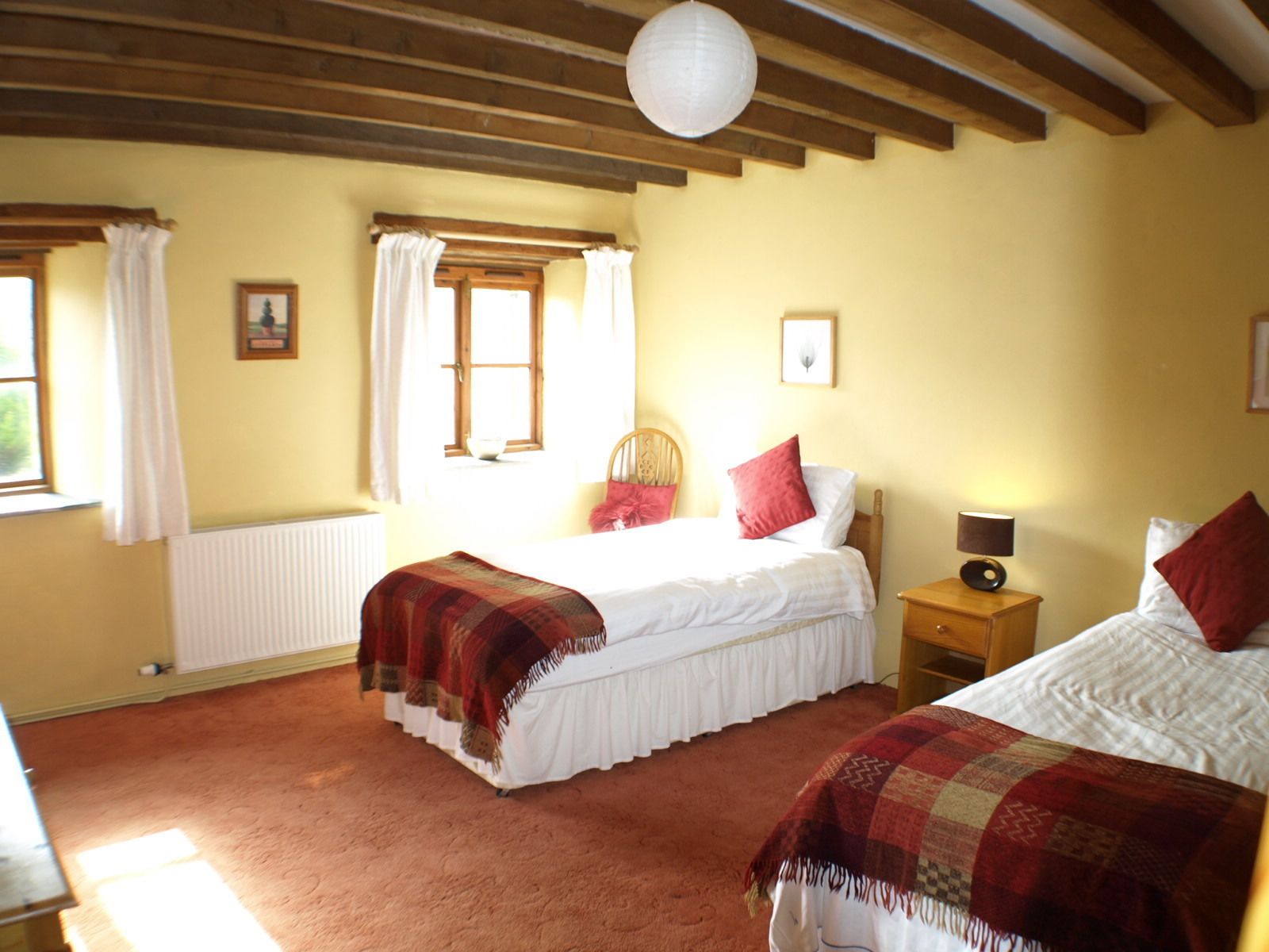 Ein gemütliches Schlafzimmer mit zwei Betten, Holzbalken an der Decke, einem Fenster mit Vorhängen und einem Nachttisch mit Lampe.