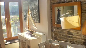 A room with a stone wall, a white sofa with cushions, a white cabinet with drawers, a sailboat model, a cityscape painting, and a window with a view of buildings. - Polean Farm Cottages (Looe)