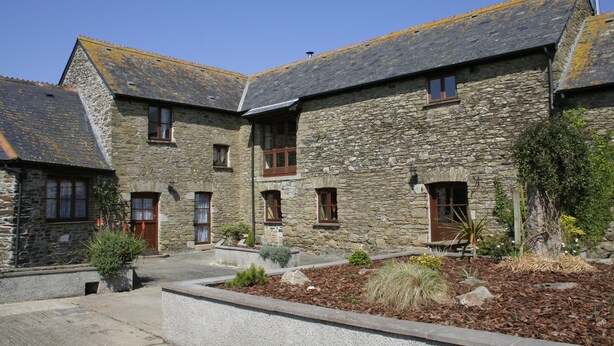 A stone building with multiple windows and doors, a courtyard with plants, and a clear blue sky. - Polean Farm Cottages (Looe)