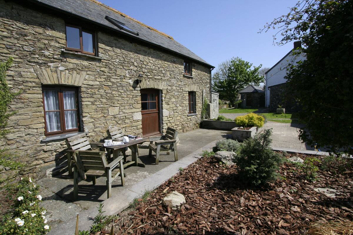 A stone building with a patio area featuring a table and chairs, surrounded by a garden with mulch and plants.