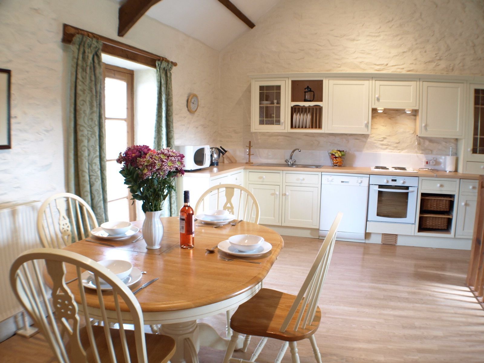 A dining area with a wooden table and white chairs, a kitchen with white cabinets and appliances, and a window with curtains.