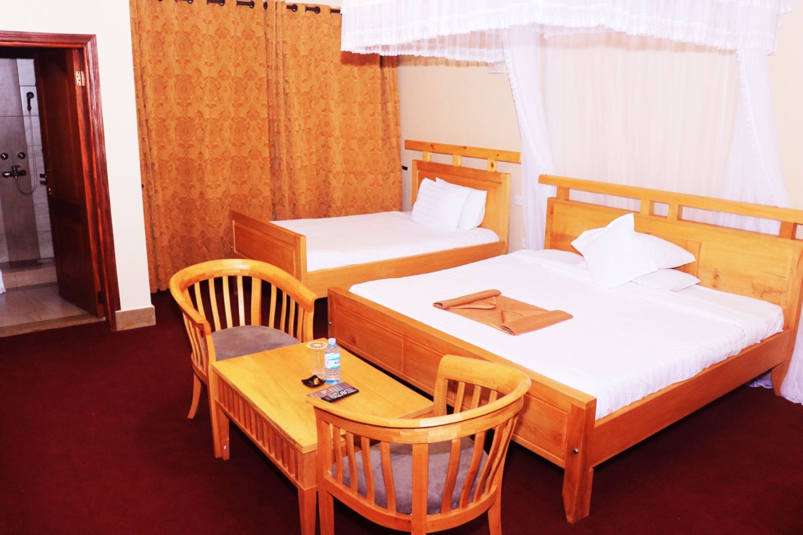 A hotel room with two wooden beds, a wooden table with chairs, a bathroom with a shower, and a door leading to another room.