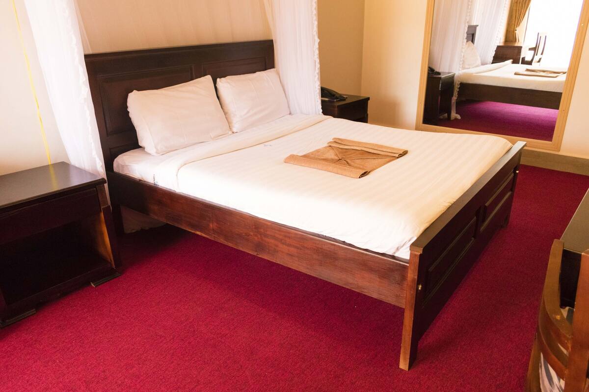 A neatly made bed with white linens and a brown folded towel, a wooden headboard, a nightstand with a phone, and a red carpeted floor.