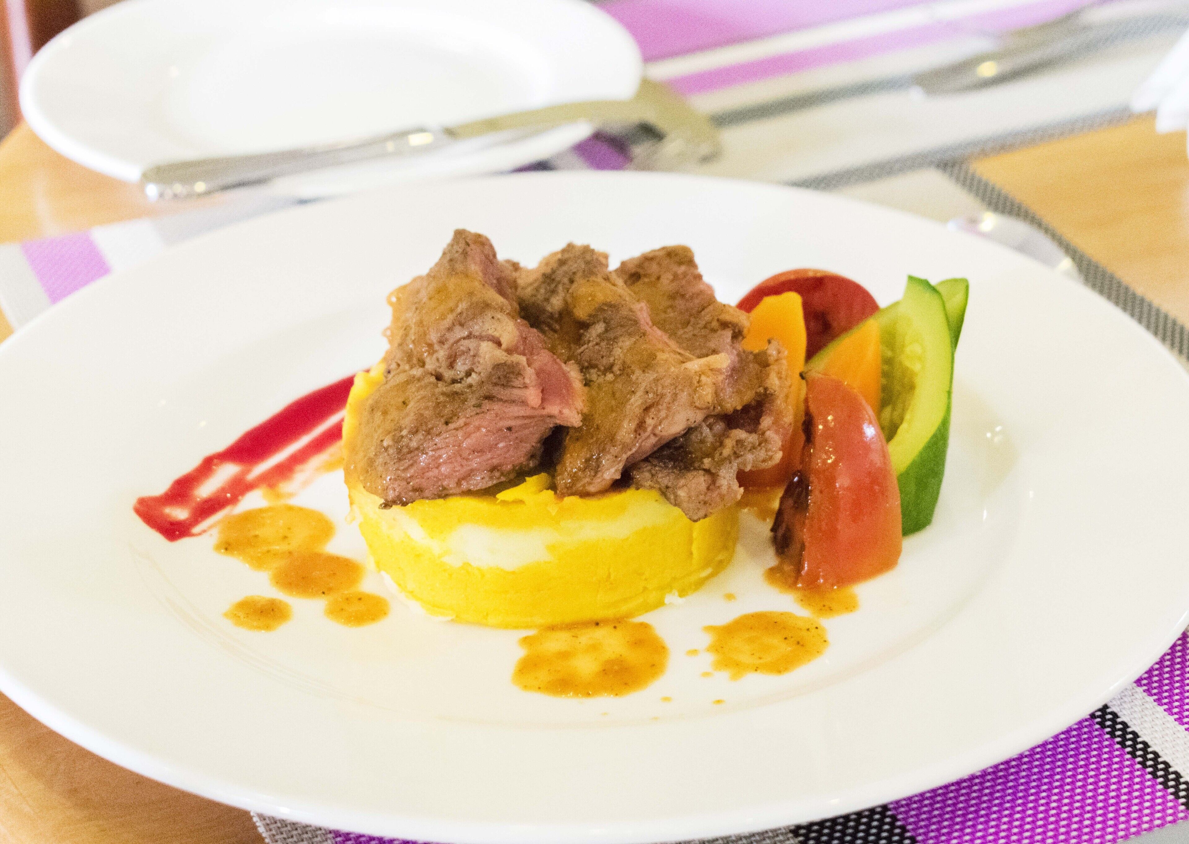 A plate with a yellow base, topped with slices of meat, surrounded by vegetables and sauce.
