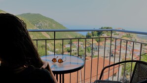 A balcony with a table and a chair, overlooking a town with red-roofed buildings and green hills. - The Z Point (Parga)
