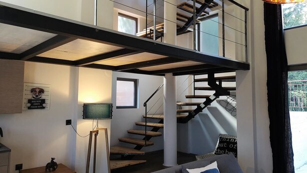 A modern loft-style living space with a staircase connecting two floors, a small table with a lamp, and a couch with cushions. - The Z Point (Parga)