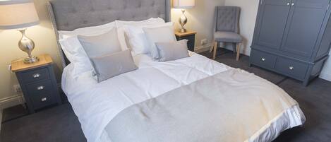 Una cama bien hecha con sábanas blancas y dos almohadas, flanqueada por mesitas de noche con lámparas, un sillón tapizado en gris y un armario gris.