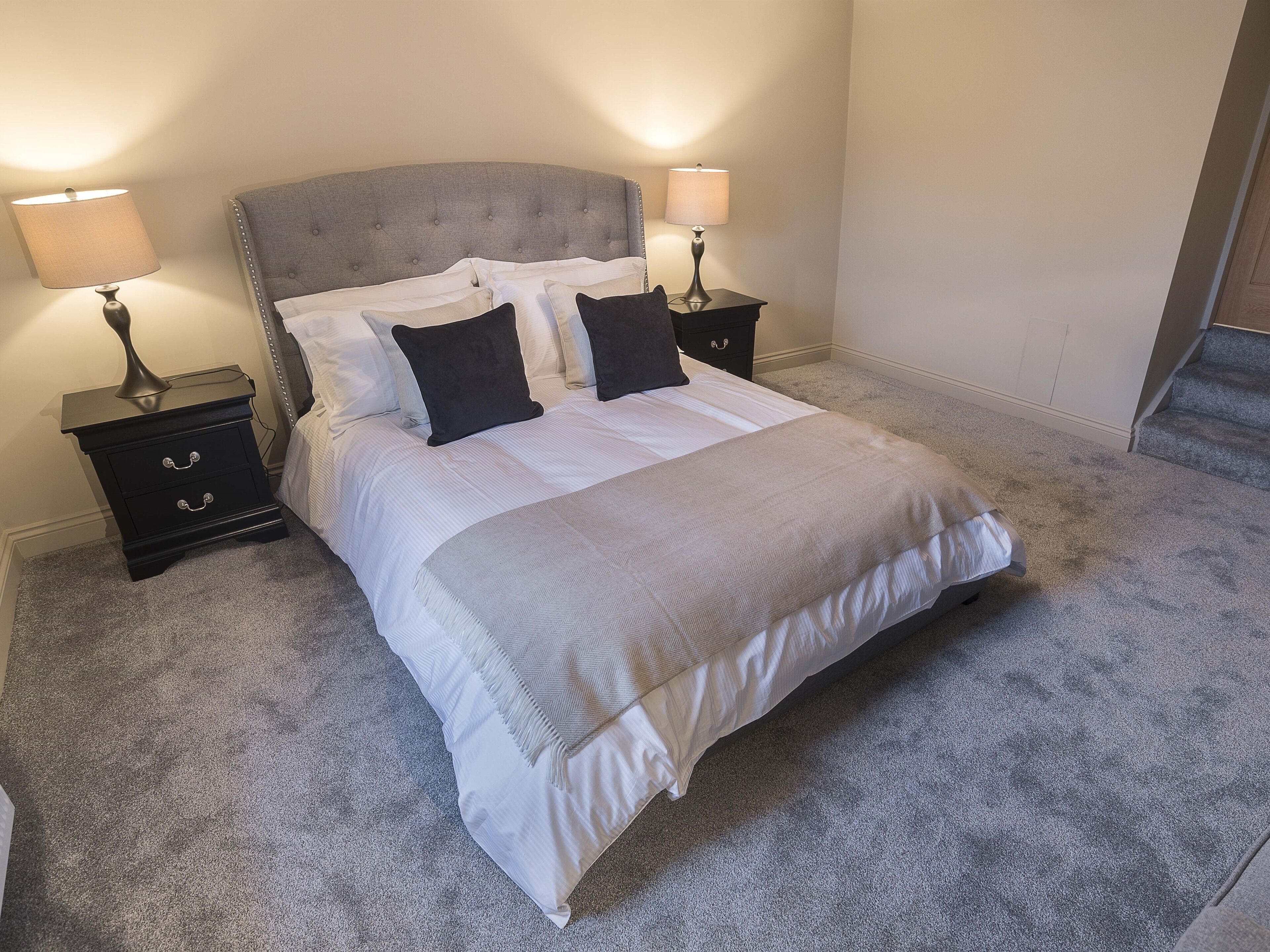 A neatly made bed with a grey headboard, two bedside tables with lamps, and a grey carpeted floor.