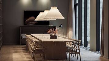 A modern dining area with a long wooden table, multiple chairs, hanging pendant lights, and a vase with flowers.