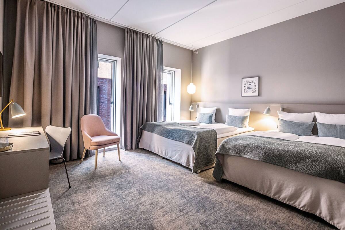 Una habitación de hotel con cama, escritorio, silla y ventana con cortinas.