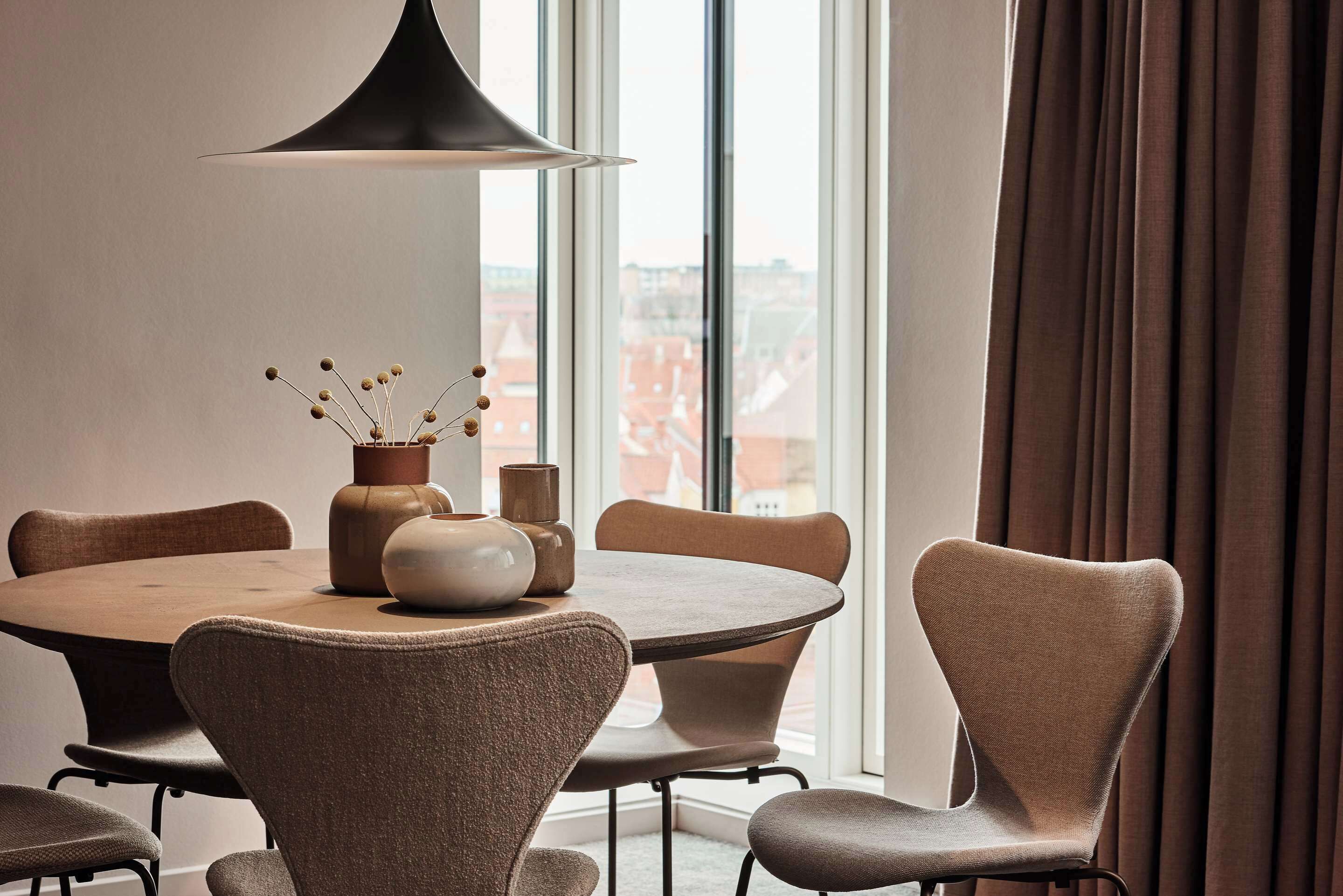 A dining area with a round table, six chairs, a modern pendant light, and a view of buildings through large windows.