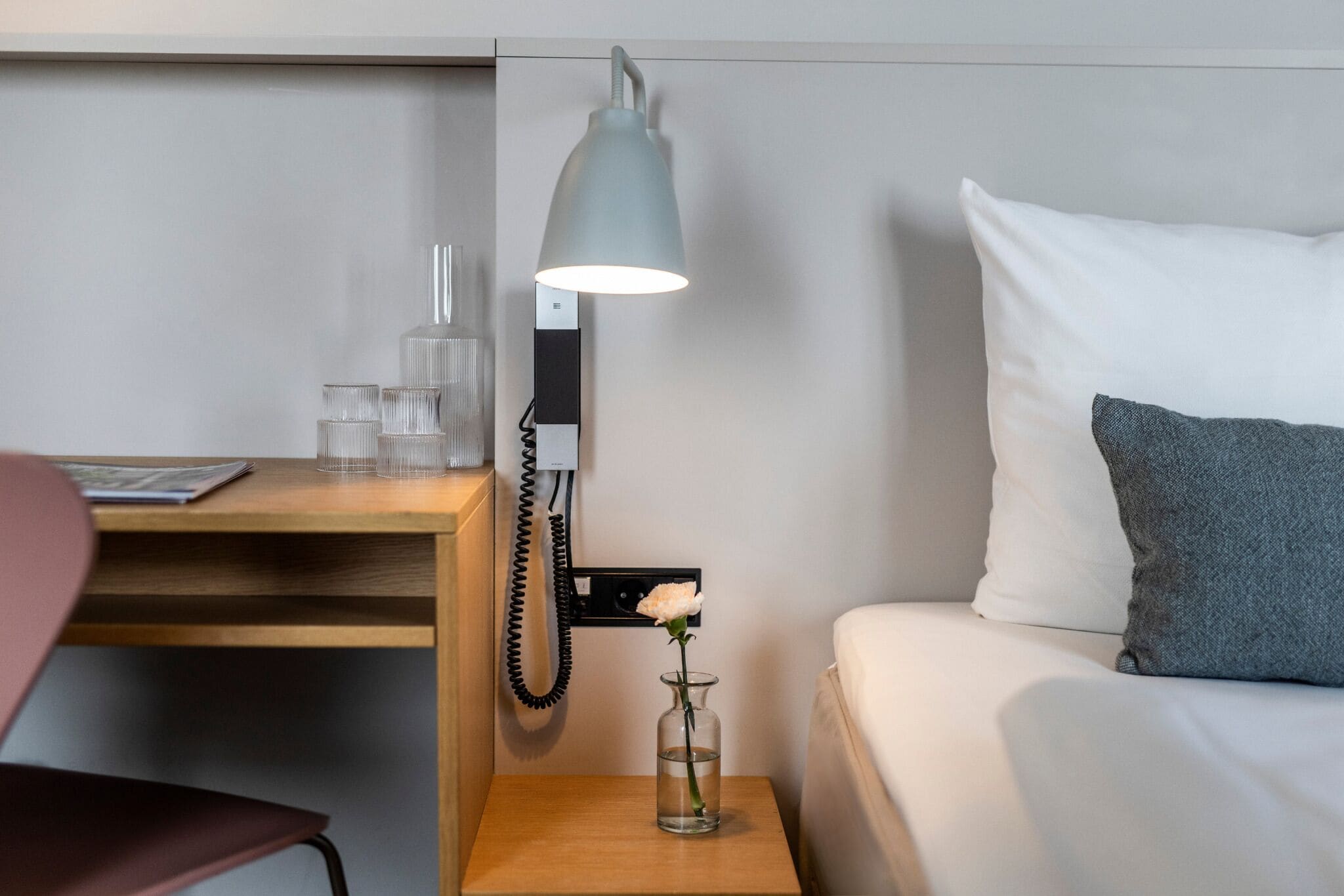 Hotellihuone, jossa on yöpöytä, lamppu, maljakko, jossa on yksi kukka, sänky tyynyineen sekä työpöytä ja tuoli.