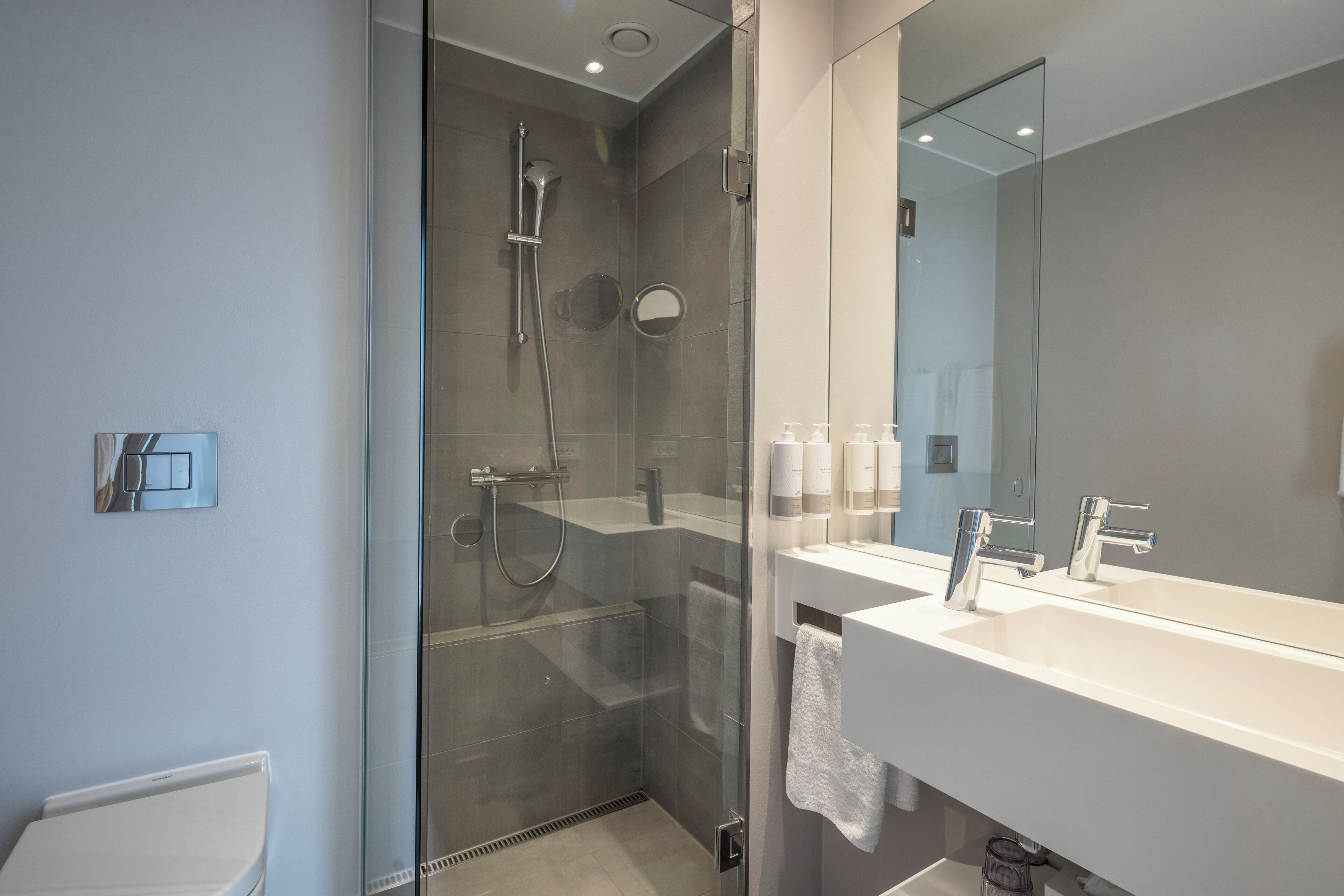 Une salle de bain moderne dotée d’une douche à parois de verre, d’un lavabo blanc avec deux robinets et d’un grand miroir placé au-dessus du lavabo.