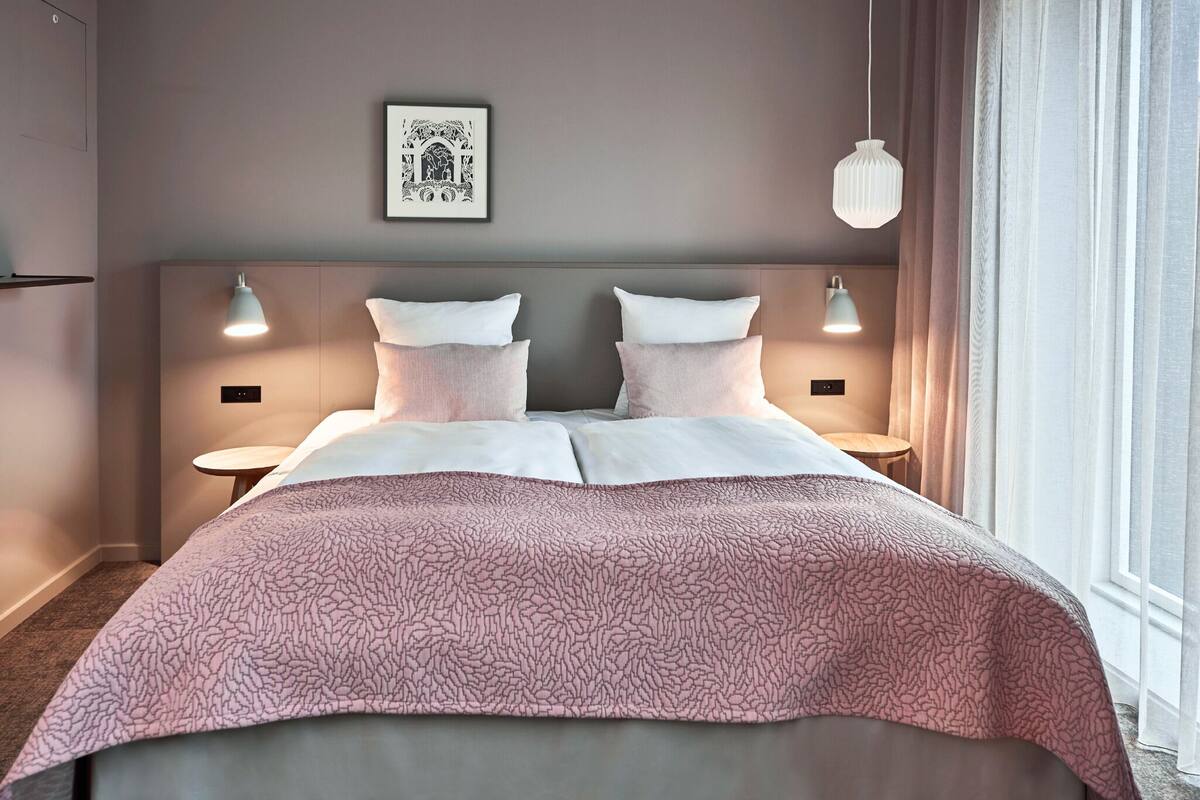 Habitación de hotel con una cama grande, dos mesitas de noche, cabecera con lámparas fijadas a la pared, un cuadro enmarcado y una ventana con cortinas.