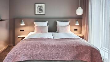 Habitación de hotel con una cama grande, dos mesitas de noche, cabecera con lámparas fijadas a la pared, un cuadro enmarcado y una ventana con cortinas.