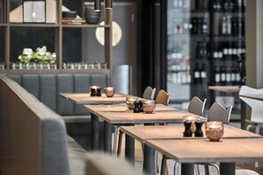 A modern restaurant interior with long tables, cushioned seating, and a bar area.