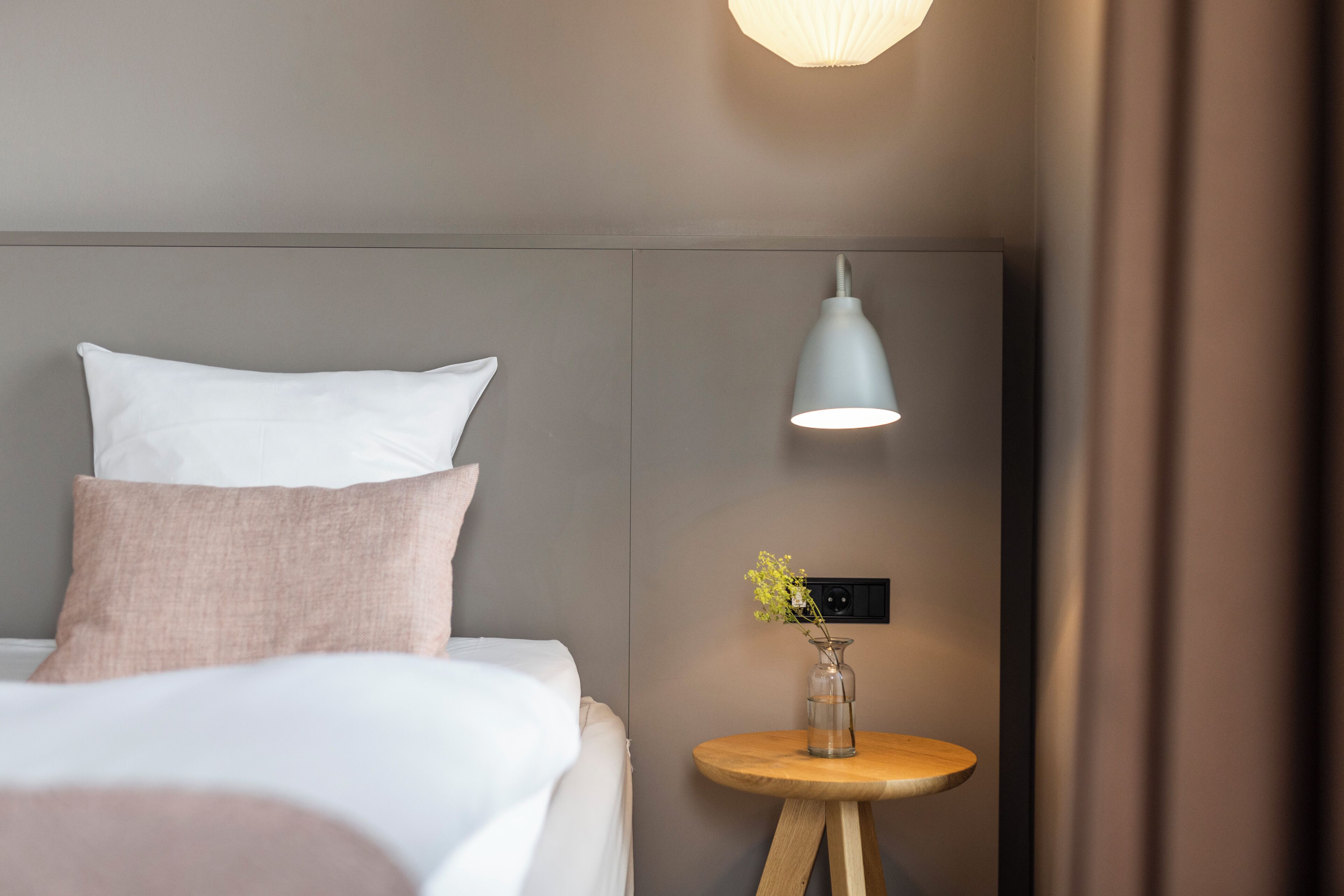 A neatly made bed with pillows, a bedside table with a vase, and a wall-mounted lamp.