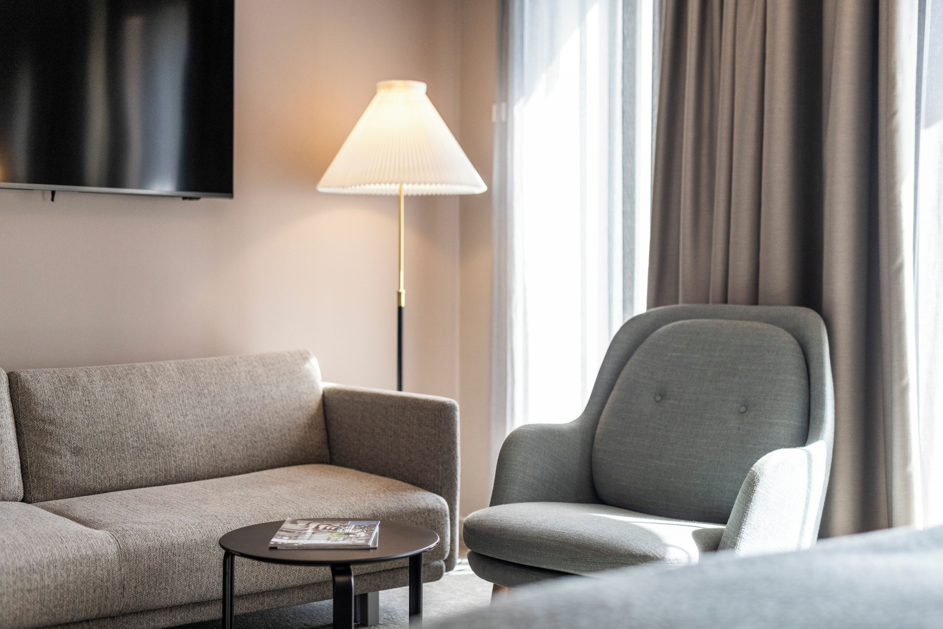 Habitación de hotel con sofá, sillón, mesita, lámpara y televisor montado en la pared.