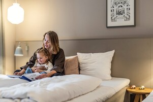Una cama con ropa de cama blanca, una mesita de noche con dos coches de juguete, una lámpara de pared y un cuadro enmarcado sobre la cama.