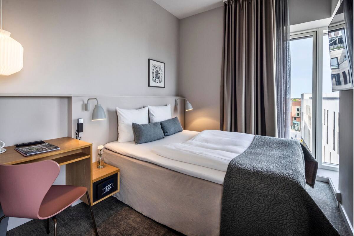 Habitación de hotel con cama, escritorio, silla, mesita de noche, lámpara, un cuadro en la pared y una ventana con cortinas.