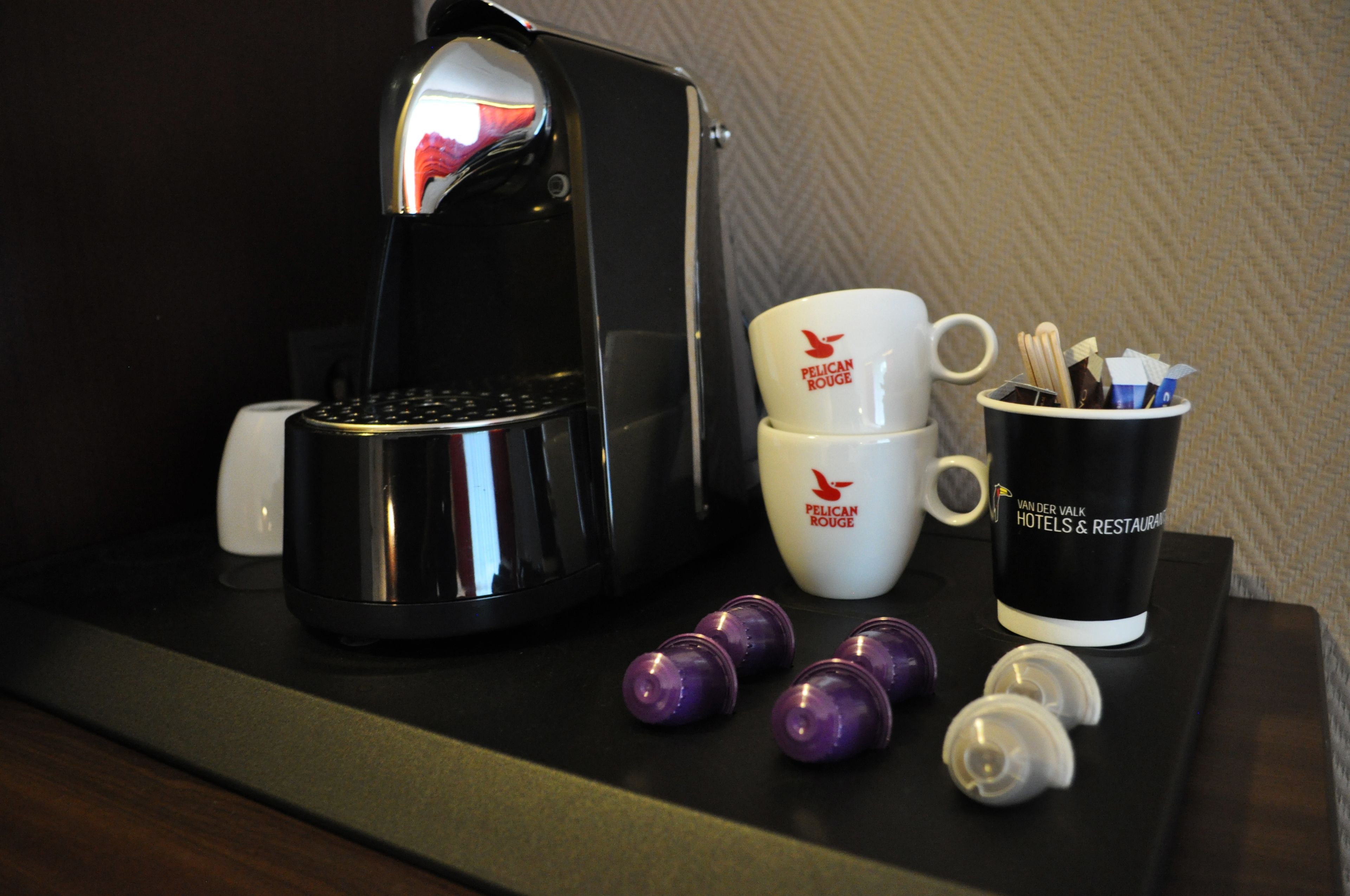 A coffee maker, two stacked cups with a logo, a mug with utensils, and two empty coffee pods on a tray.