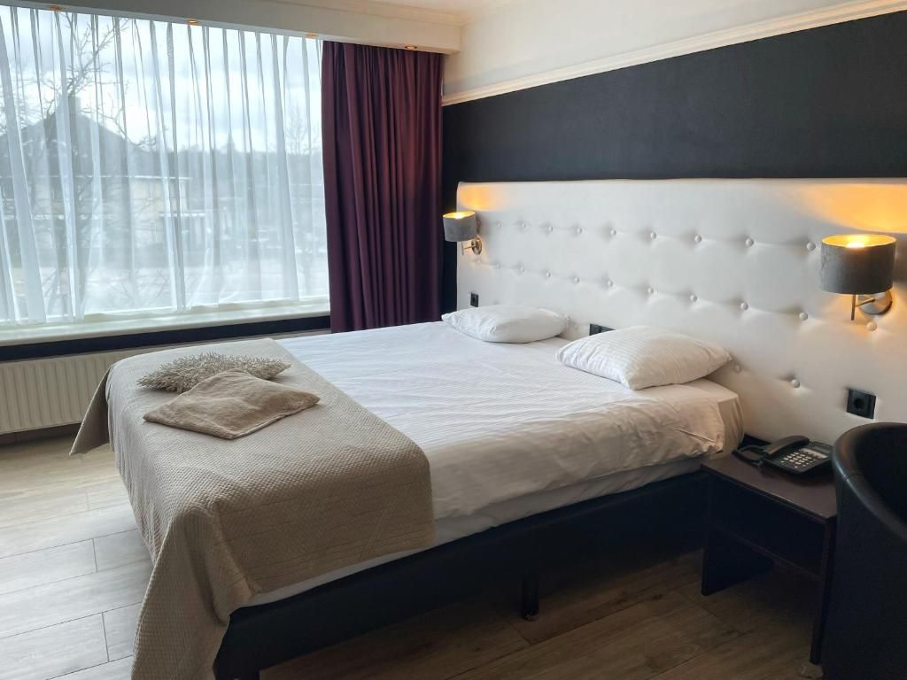 Et hotelværelse med en stor seng, et sengebord med lampe og udsigt ud af et vindue med gardiner.