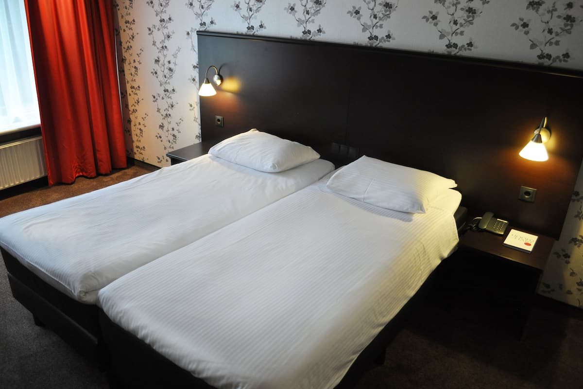 Un lit double avec du linge de lit blanc, deux tables de chevet avec des lampes et une fenêtre avec des rideaux rouges.