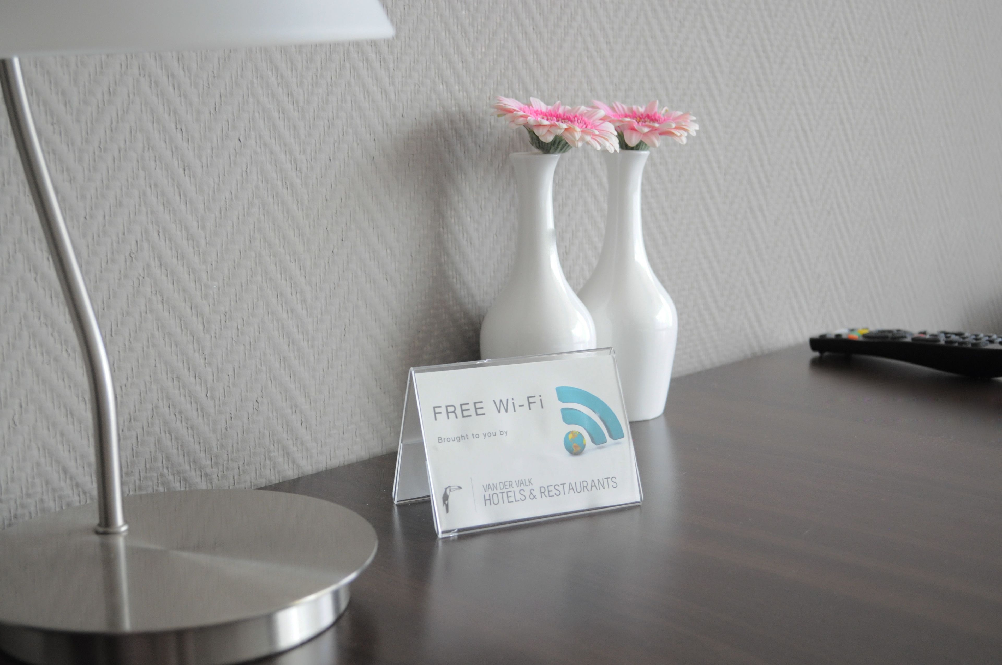 Habitación de hotel con un cartel que indica Wi-Fi gratuito, un control remoto y dos jarrones blancos con flores rosadas sobre la mesa.