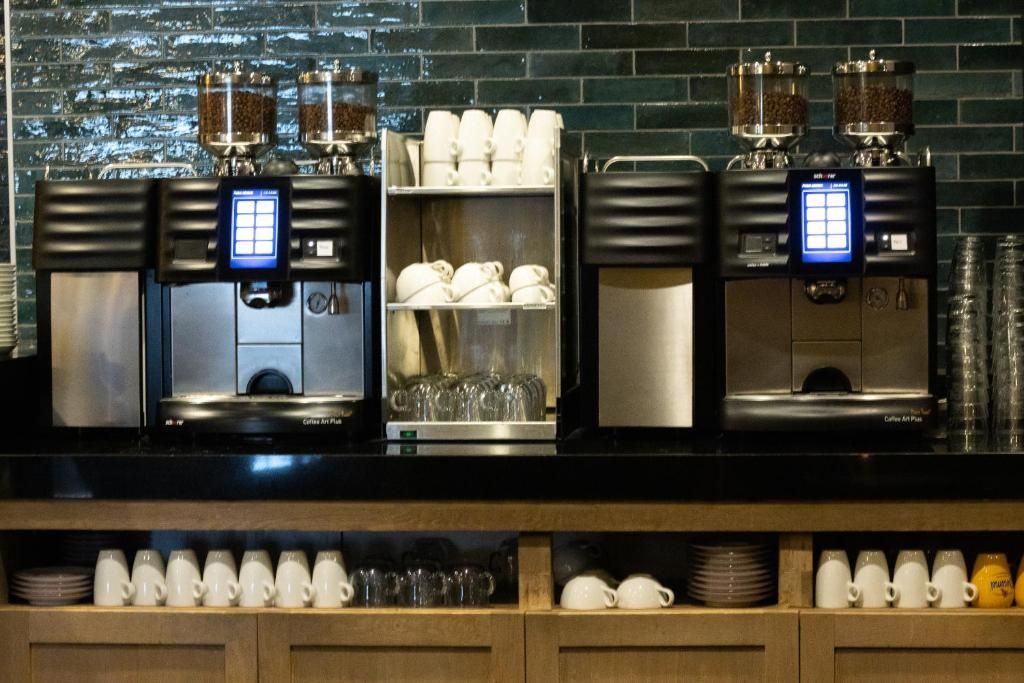 A coffee station with multiple brewing machines, a shelf with cups and saucers, and a brick wall background.