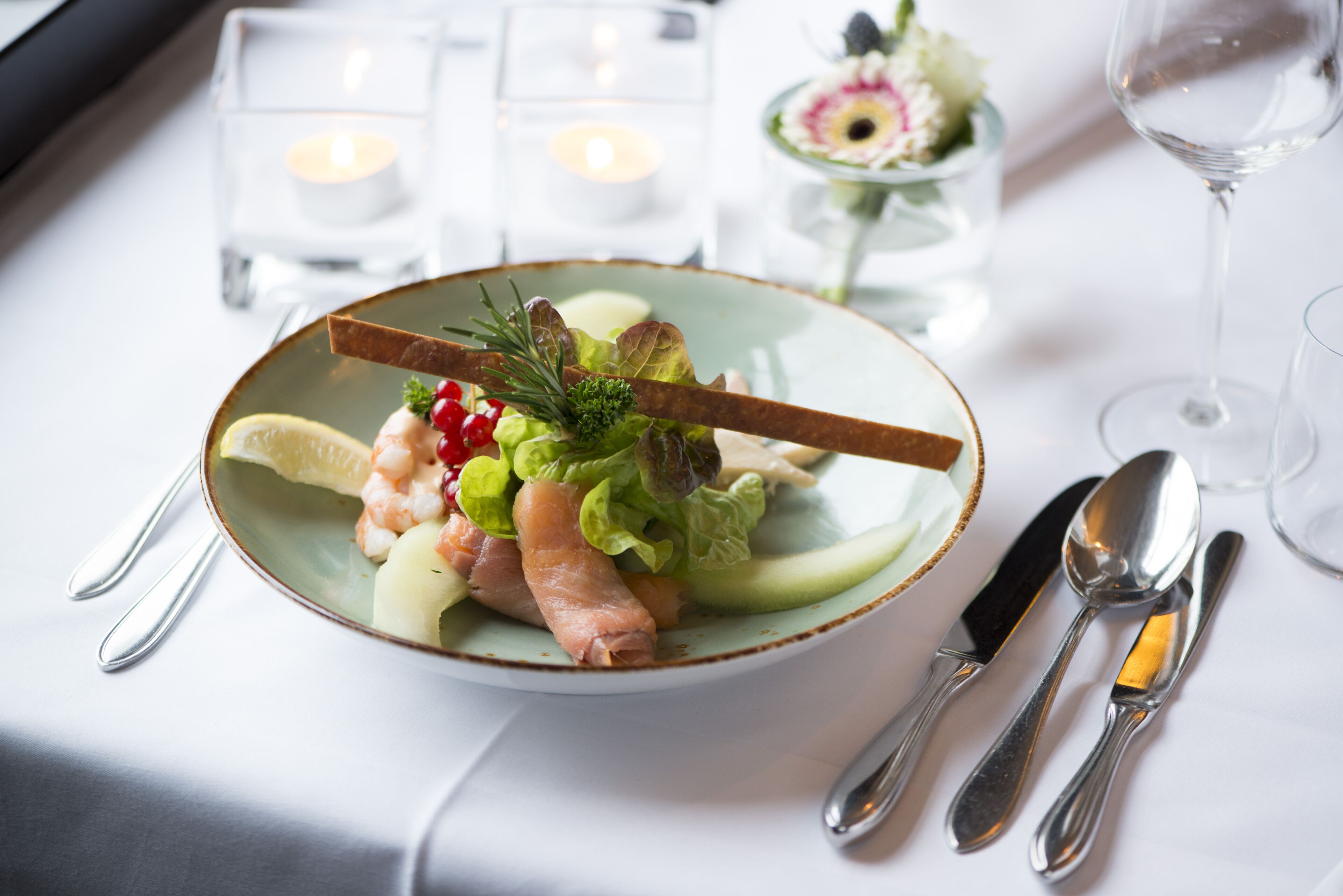 A plate of food with a lemon wedge, shrimp, and greens, accompanied by cutlery and candles on a table.