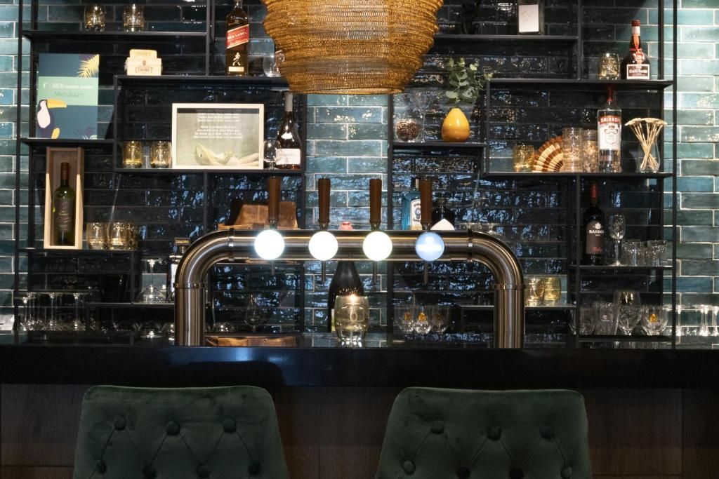 Eine Bar mit einer Wand aus schwarzen Ziegelsteinen, Regalen mit Flaschen und Gläsern sowie zwei grünen Polstersesseln.